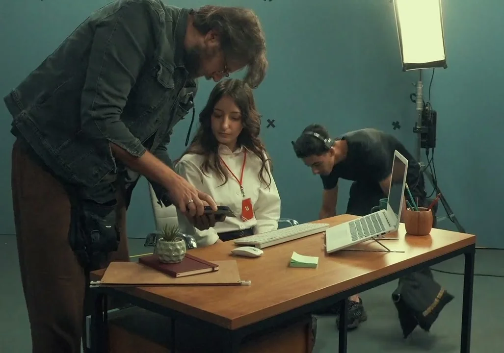 Diretor mostra o celular à atriz sentada à mesa de escritório com laptop e teclado, enquanto um técnico ajusta o set ao fundo.