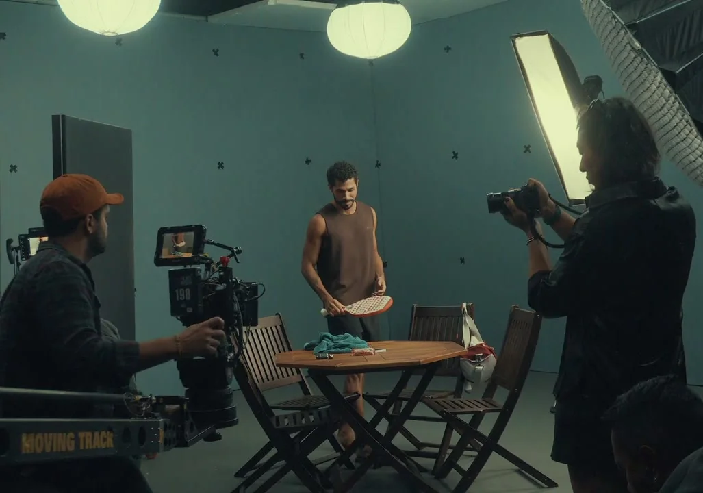 Set da cena da mesa: operador de câmera em trilho e fotógrafo enquadram o ator junto a uma mesa de madeira real no estúdio.